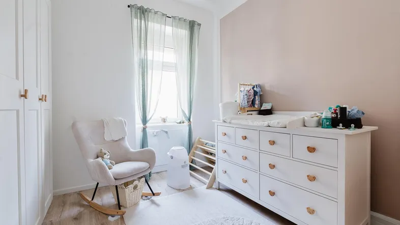 Helles Kinderzimmer mit Wickelkommode, Schaukelstuhl und hellem Holzfußboden.