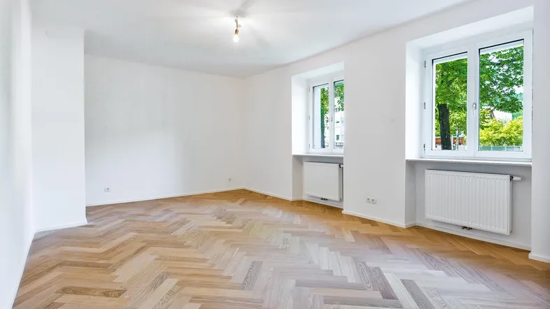 Helles Zimmer mit Fischgrätparkettboden, weißen Wänden und Blick ins Grüne durch zwei Fenster.