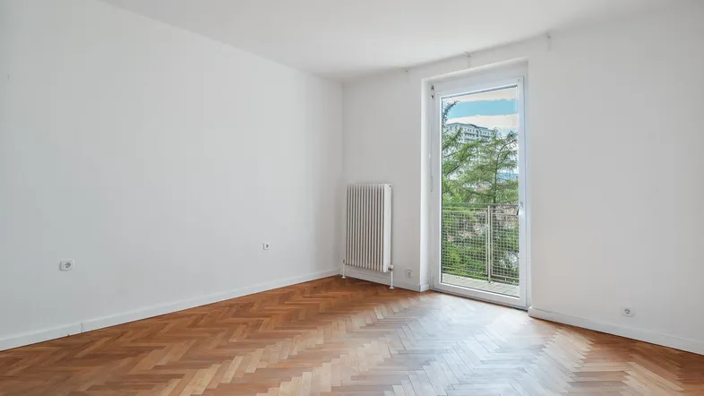 Helles Zimmer mit Fischgrätparkettboden und Zugang zu einem kleinen Balkon.