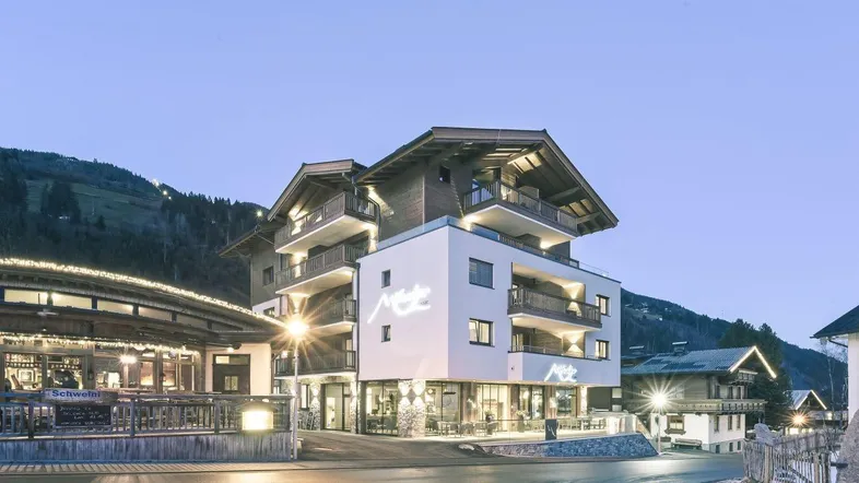Modernes Gebäude mit Holz- und Putzfassade, beleuchtet in der Abenddämmerung, umgeben von Bergen.
