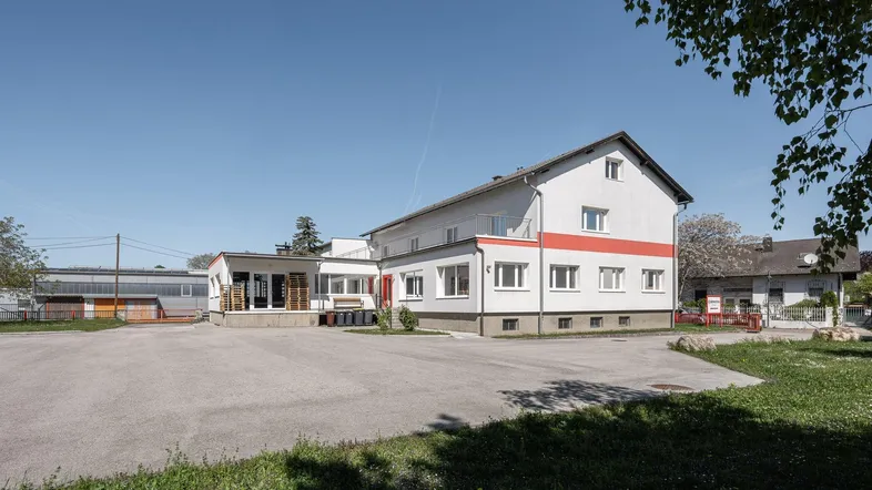Gebäudeansicht mit großem Vorplatz und umliegender Grünfläche unter blauem Himmel.