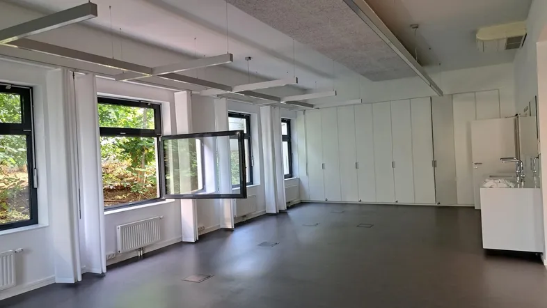 Helles, modernes Mehrzweckzimmer mit großen Fenstern, viel Stauraum und Waschbecken. Die Decke ist mit Akustikplatten ausgestattet.