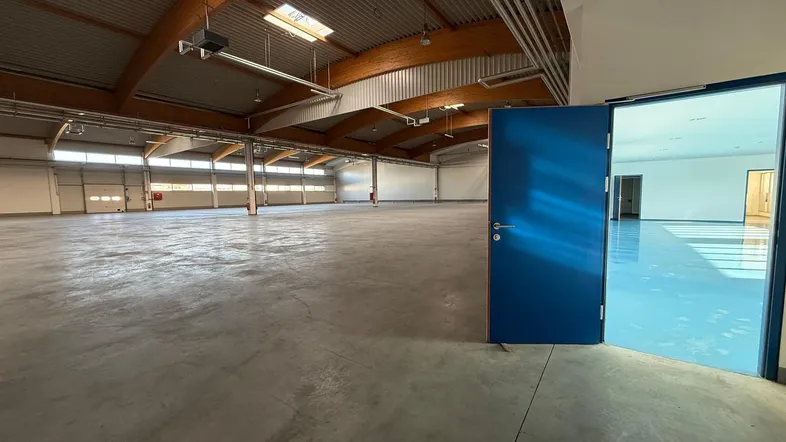 Geräumige Lagerhalle mit offenem blauen Tor, das zu einem weiteren hellen Raum führt, mit Betonboden.