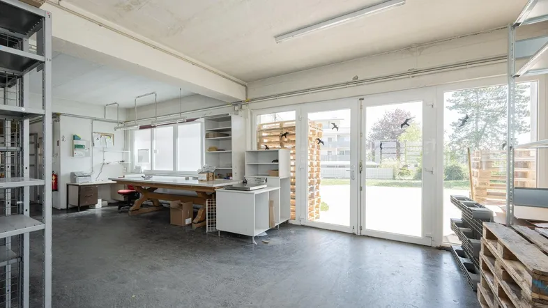 Geräumiger Werkstatt- oder Lagerraum mit großen Fenstern und direktem Zugang zum Außenbereich, ausgestattet mit Regalen und Arbeitsflächen.
