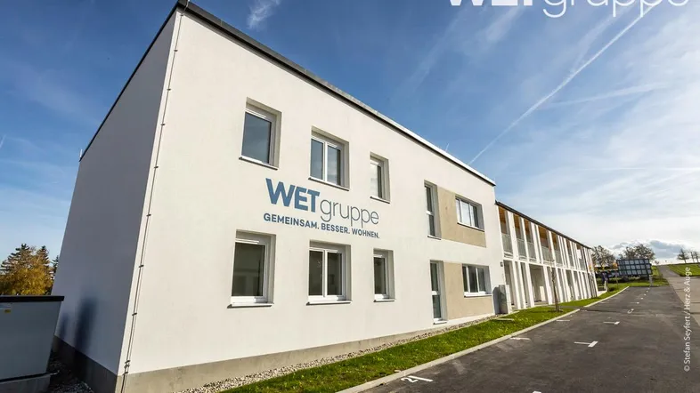 Modernes Mehrfamilienhaus mit weißer Fassade und Parkplätzen unter blauem Himmel.