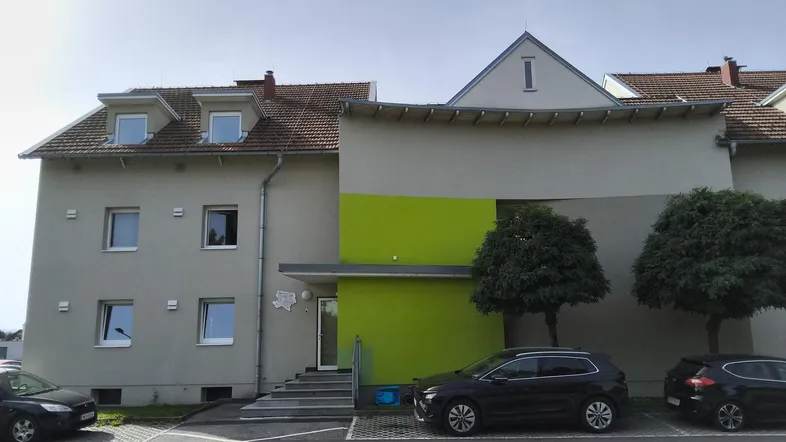 Modernes Mehrfamilienhaus mit hellgrauer Fassade, grünem Akzent und Parkplätzen davor.