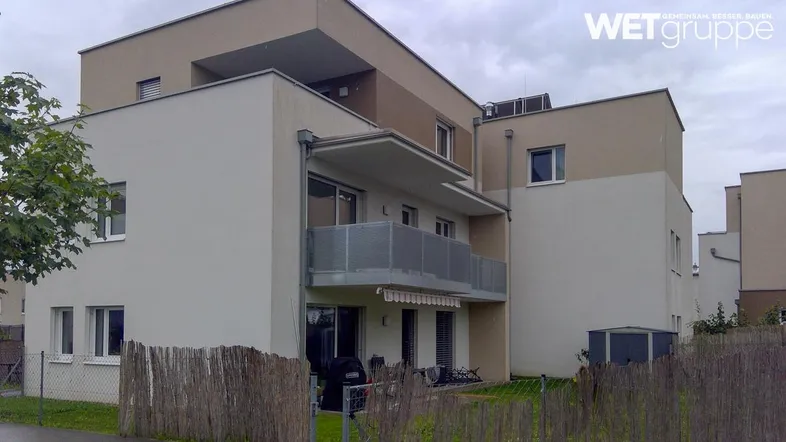 Modernes Mehrfamilienhaus mit Balkon, Gartenbereich und Jalousien an den Fenstern unter bewölktem Himmel.