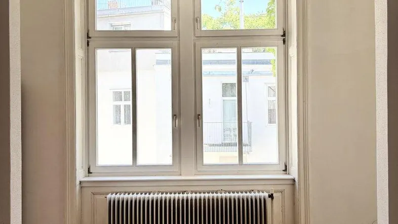 Helles Zimmer mit Fischgrätparkett, großem Fenster und Heizkörper, durch einen Bogenrahmen gesehen.
