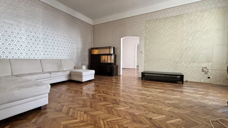 Geräumiges Wohnzimmer mit klassischem Fischgrätparkett, gemusterter Tapete und einem großen Ecksofa.
