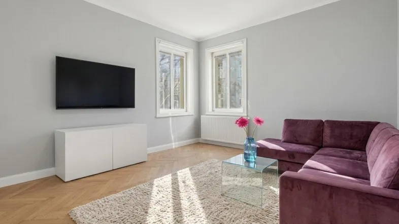 Helles Wohnzimmer mit modernem Sofa, Fernseher und Fischgrätparkettboden, durchflutet von Tageslicht.