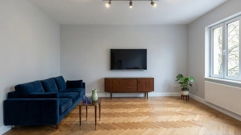 Helles Wohnzimmer mit modernem blauen Sofa, Holzboden im Fischgrätmuster und einem Fenster mit Blick ins Grüne.