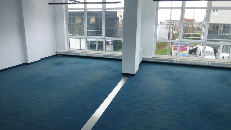 Geräumiger, leerer Büroraum mit großen Fenstern und blauem Teppichboden, bereit zur individuellen Gestaltung.
