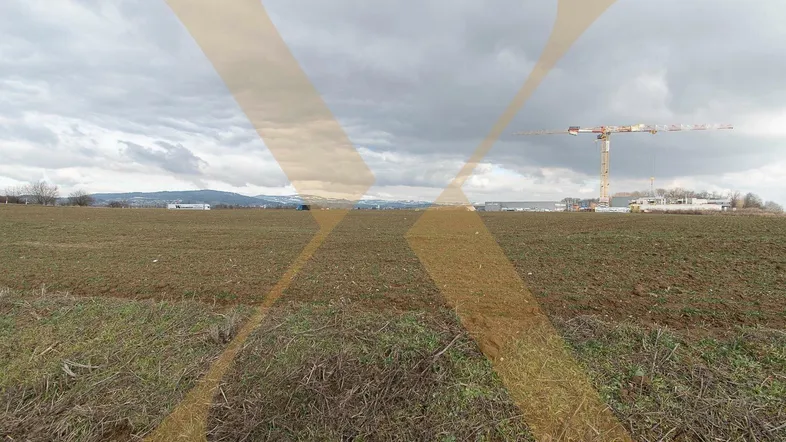 Weites Feld mit Blick auf entfernte Gebäude, einen Baukran und eine Bergkette unter bewölktem Himmel.