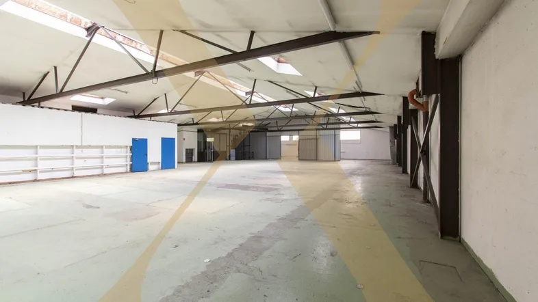 Große, leere Lagerhalle mit hohen Decken, sichtbaren Stahlträgern und Oberlichtern für Tageslicht.