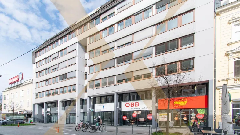 Modernes Mehrzweckgebäude mit zahlreichen Fenstern und Geschäften im Erdgeschoss, darunter ein Supermarkt und ein ÖBB-Büro.