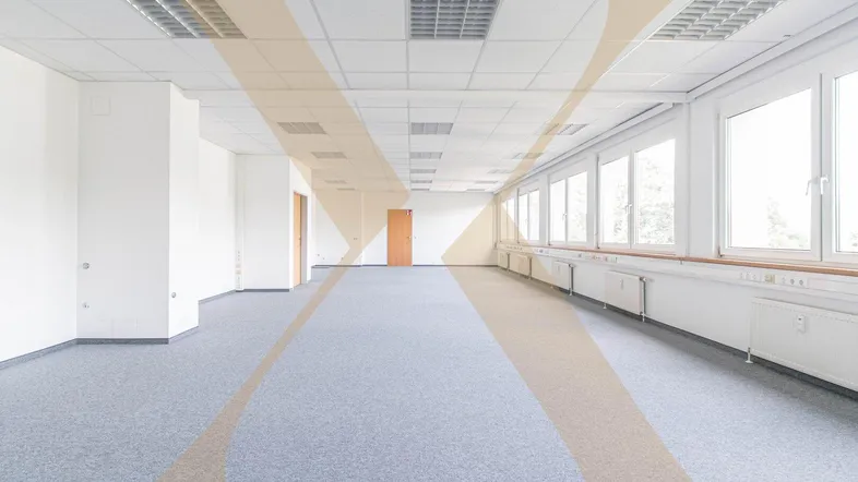 Geräumiges, leeres Büro mit grauem Teppichboden, weißen Wänden und vielen Fenstern für natürliches Licht.