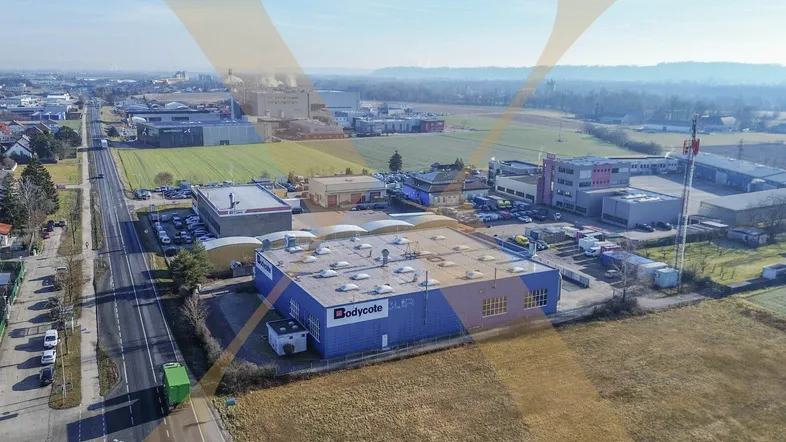 Luftaufnahme eines modernen Industriegebäudes mit blauer Fassade, umgeben von Feldern und einem Gewerbegebiet.