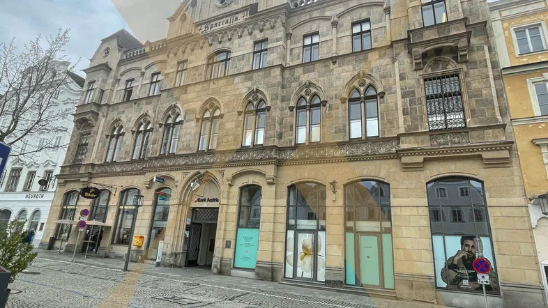 Historisches Gebäude mit reich verzierter Fassade und großen Fenstern im Erdgeschoss, gelegen an einer gepflasterten Straße.
