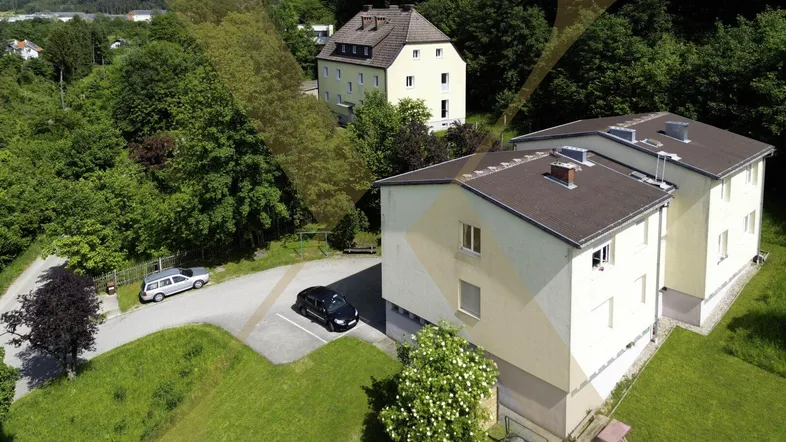 Luftaufnahme von zwei Mehrfamilienhäusern mit Parkplätzen und viel Grün in hügeliger Umgebung.