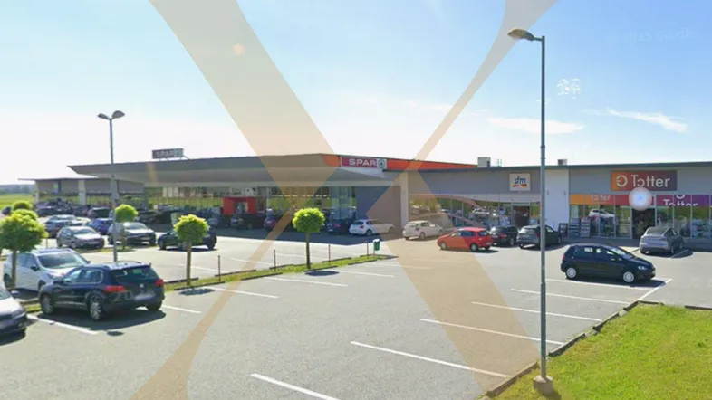 Weitläufiger Parkplatz vor einem modernen Einkaufszentrum mit verschiedenen Geschäften unter blauem Himmel.