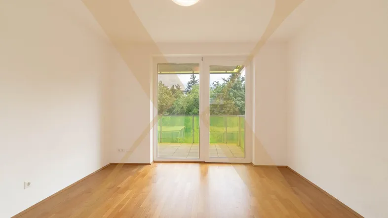 Helles Zimmer mit Holzboden und Zugang zu einem Balkon mit Blick ins Grüne.