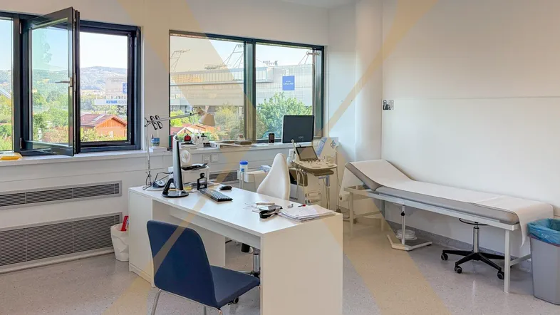 Helles Behandlungszimmer mit großen Fenstern, Schreibtisch und Untersuchungsliege, ideal für eine Arztpraxis.