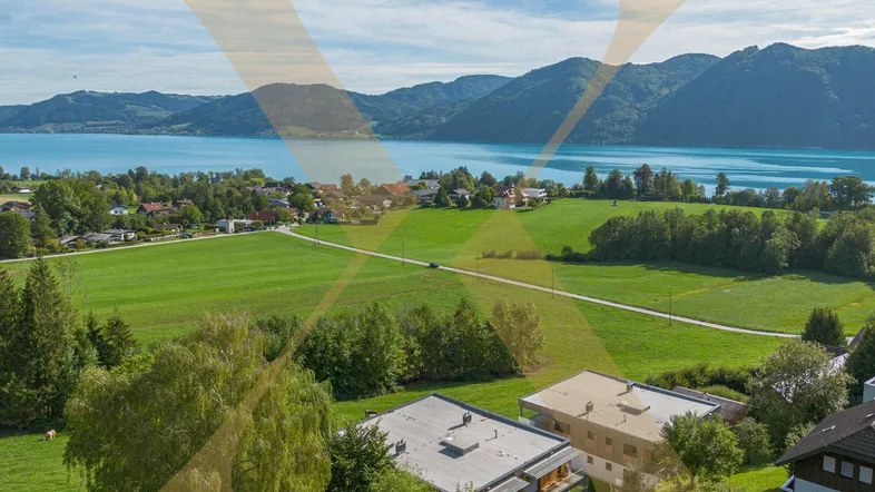 Panoramablick auf den See, umgeben von grünen Hügeln und Bergen, mit einigen Häusern in der Landschaft.