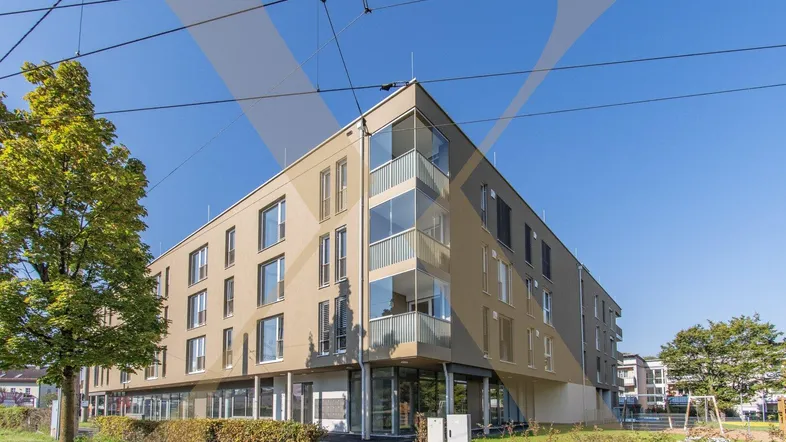 Modernes Mehrfamilienhaus mit beiger Fassade und großen Eckfenstern an einer belebten Straße gelegen.