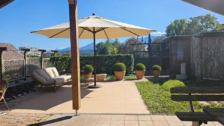 Große gepflasterte Terrasse mit Sonnenschirm und Gartenmöbeln, umgeben von Grün und mit Bergblick.