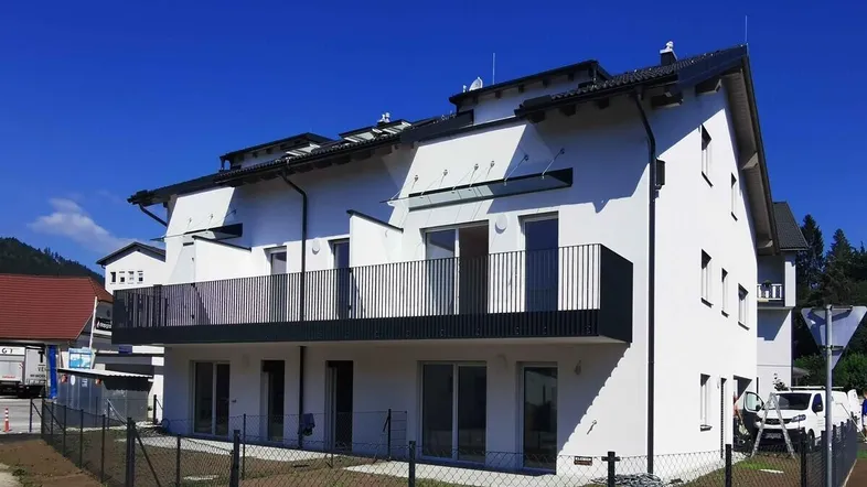 Modernes Mehrfamilienhaus mit weißer Fassade, schwarzen Balkonen und Satteldach unter blauem Himmel.