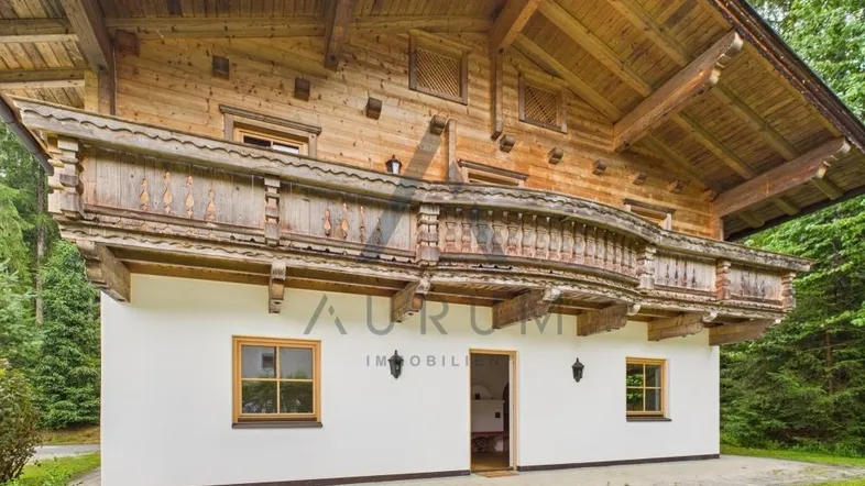 Traditionelles Holzchalet mit weißer Fassade und geschnitztem Holzbalkon, umgeben von grüner Natur.