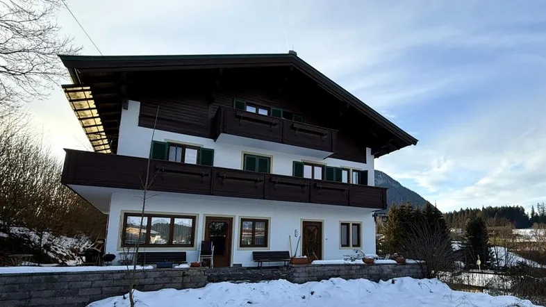 Traditionelles Haus im alpinen Stil mit Holzbalkonen und verschneiter Umgebung vor Bergkulisse.