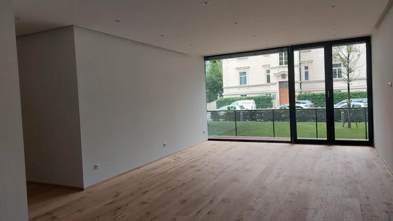 Helles, leeres Zimmer mit Holzboden und großen Fenstern, die auf die Straße blicken.