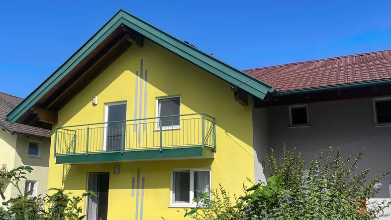 Gelbe Hausfassade mit grünem Dach und Balkon unter blauem Himmel.