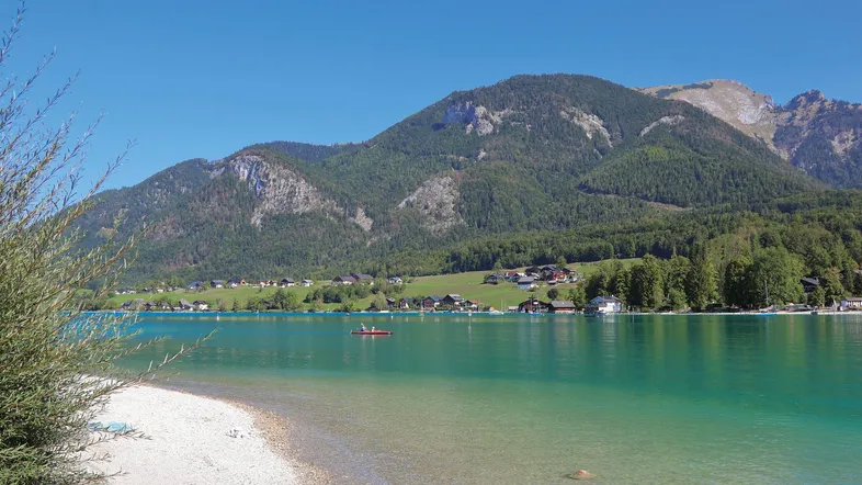 Malerischer See mit klarem türkisfarbenem Wasser, umgeben von grünen Bergen und Häusern am Ufer.