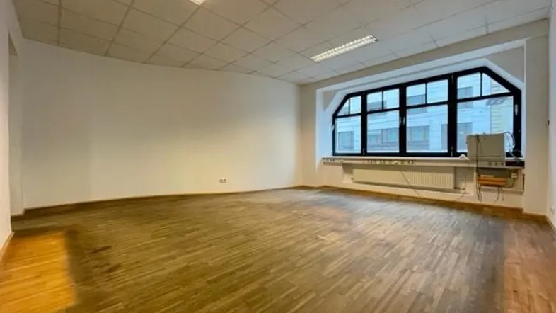 Geräumiger, leerer Büroraum mit Holzfußboden und großen Fenstern, die viel Tageslicht hereinlassen.