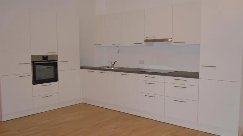 Moderne, leere Küche mit weißen Schränken, integriertem Backofen und hellem Holzfußboden.
