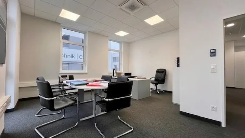 Modernes Büro mit Konferenztisch, ergonomischen Stühlen und Teppichboden, gut beleuchtet durch Deckenleuchten und Fenster.