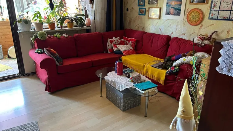 Gemütliches Wohnzimmer mit rotem Ecksofa, vielen Pflanzen und Zugang zu einem Balkon.