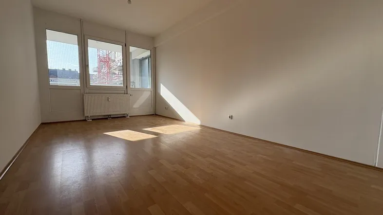 Helles, leeres Zimmer mit Holzfußboden und großen Fenstern, das viel Tageslicht hereinlässt.