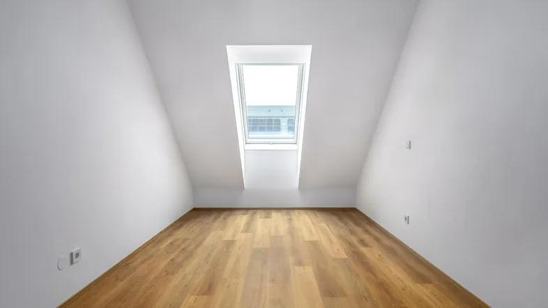 Heller Raum im Dachgeschoss mit schrägen Wänden, Dachfenster und Holzdielenboden.