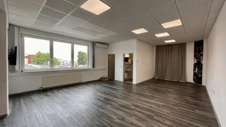 Geräumiger Büroraum mit dunklem Laminatboden, großen Fenstern und abgehängter Decke mit integrierter Beleuchtung.