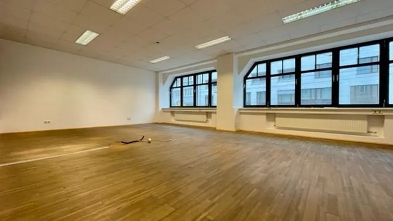 Großer, leerer Büroraum mit Holzparkettboden und hohen Fenstern, die viel Tageslicht hereinlassen.