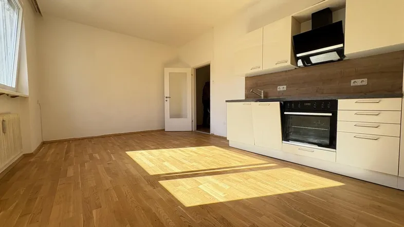 Helles Zimmer mit Holzboden und integrierter moderner Küchenzeile, durchflutet von Sonnenlicht.