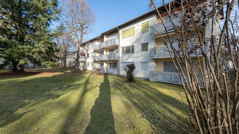 Mehrfamilienhaus mit Balkonen und großzügiger Grünfläche im Vordergrund unter blauem Himmel.