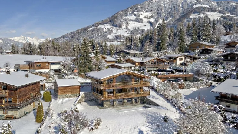 Luftaufnahme eines luxuriösen Holzchalets in einer verschneiten Berglandschaft unter blauem Himmel.