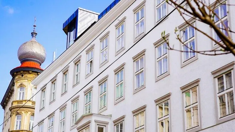 Fassade eines weißen Gebäudes mit vielen Fenstern und Erker, neben einem historischen Gebäude mit Kuppel.