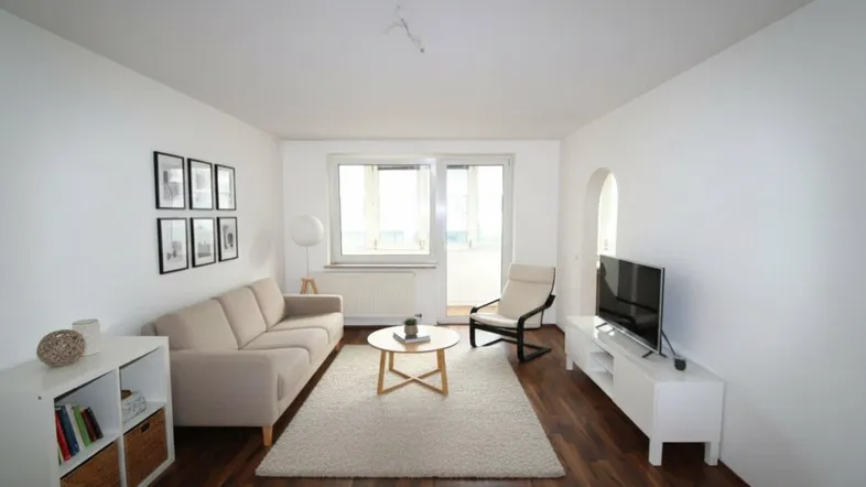 Helles Wohnzimmer mit dunklem Parkettboden, beigem Sofa und Zugang zum Balkon.