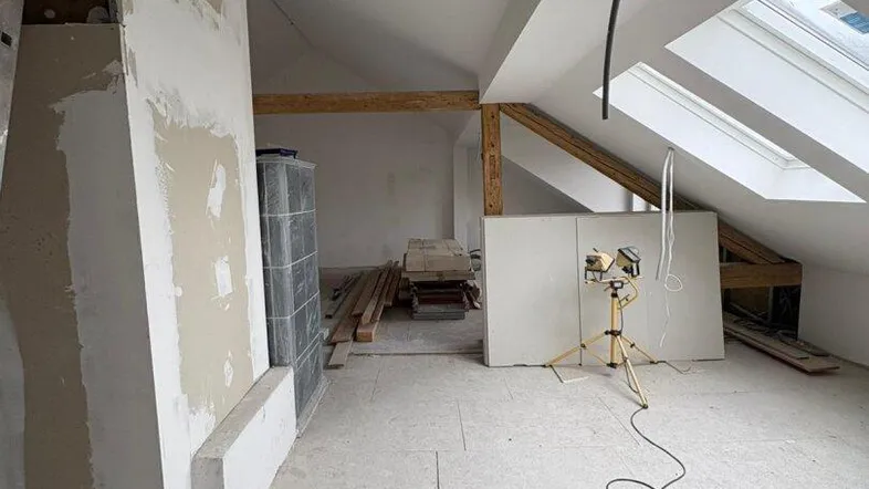 Dachgeschoss im Umbau mit sichtbaren Holzbalken, Dachfenstern und einem Kachelofen.