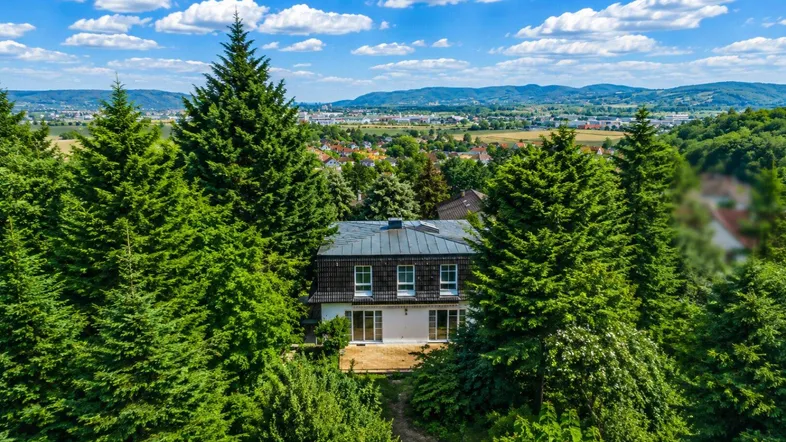 Luftaufnahme eines Hauses, umgeben von üppigem Grün und hohen Bäumen, mit Fernblick auf die Landschaft.